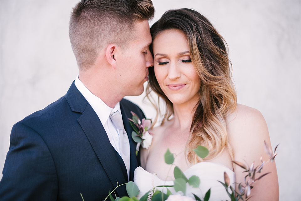 Orange county outdoor wedding at old ranch country club bride strapless ballgown with a sweetheart neckline and beaded detail with groom navy notch lapel suit with matching vest and white dress shirt with long silver tie and white floral boutonniere kissing and bride holding light pink and green floral bridal bouquet close up