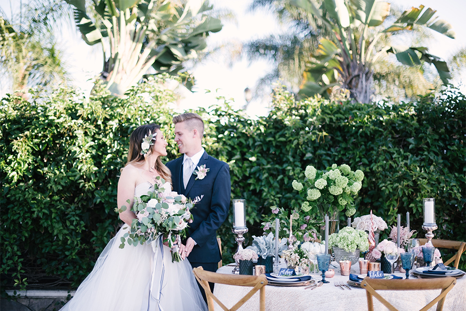 Orange county outdoor wedding at old ranch country club bride strapless ballgown with a sweetheart neckline and beaded detail with groom navy notch lapel suit with matching vest and white dress shirt with long silver tie and white floral boutonniere standing by table bride holding light pink and green floral bridal bouquet