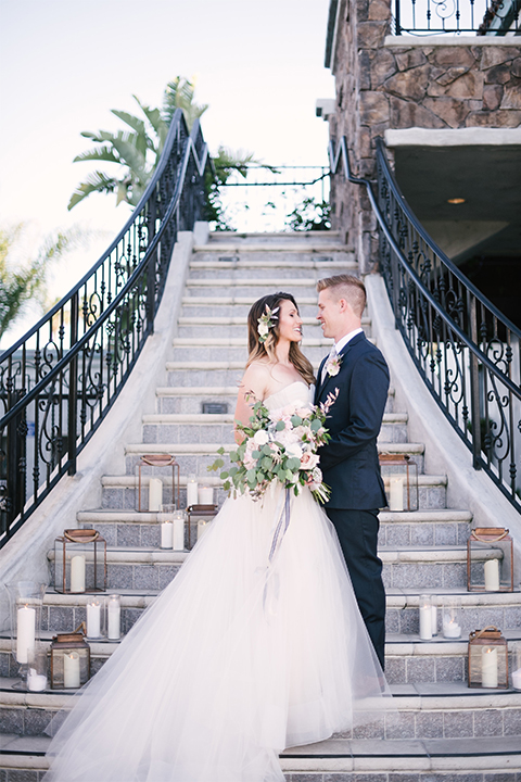 Orange county outdoor wedding at old ranch country club bride strapless ballgown with a sweetheart neckline and beaded detail with groom navy notch lapel suit with matching vest and white dress shirt with long silver tie and white floral boutonniere hugging and standing by stairs bride holding light pink and green floral bridal bouquet with candle decor