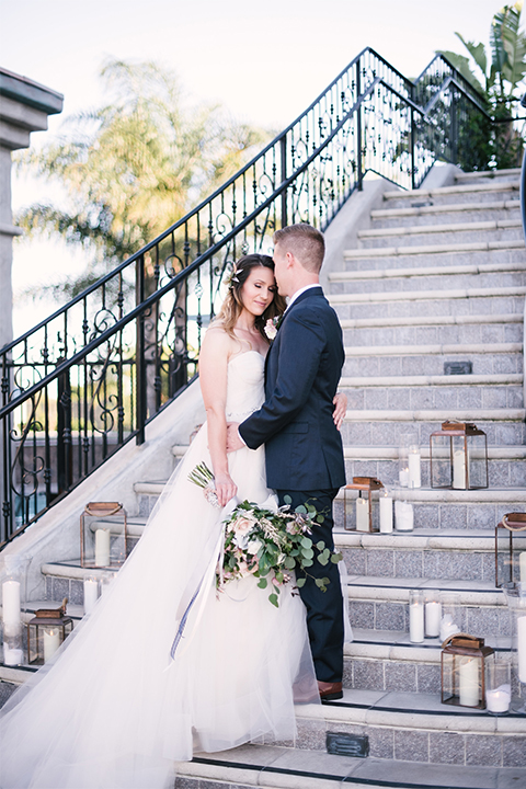 Orange county outdoor wedding at old ranch country club bride strapless ballgown with a sweetheart neckline and beaded detail with groom navy notch lapel suit with matching vest and white dress shirt with long silver tie and white floral boutonniere hugging and standing by stairs bride holding light pink and green floral bridal bouquet with candle decor