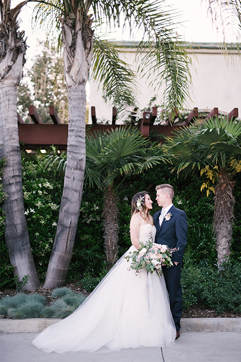 Orange county outdoor wedding at old ranch country club bride strapless ballgown with a sweetheart neckline and beaded detail with groom navy notch lapel suit with matching vest and white dress shirt with long silver tie and white floral boutonniere hugging and bride holding light pink and green floral bridal bouquet