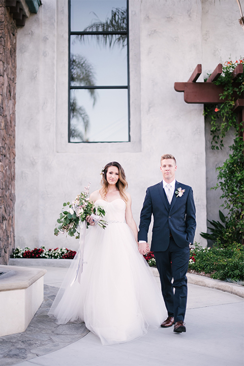 Orange county outdoor wedding at old ranch country club bride strapless ballgown with a sweetheart neckline and beaded detail with groom navy notch lapel suit with matching vest and white dress shirt with long silver tie and white floral boutonniere standing and holding hands bride holding light pink and green floral bridal bouquet