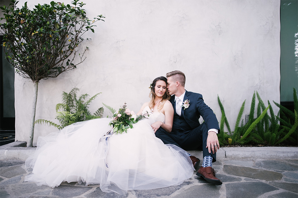 Orange county outdoor wedding at old ranch country club bride strapless ballgown with a sweetheart neckline and beaded detail with groom navy notch lapel suit with matching vest and white dress shirt with long silver tie and white floral boutonniere sitting and hugging bride holding light pink and green floral bridal bouquet