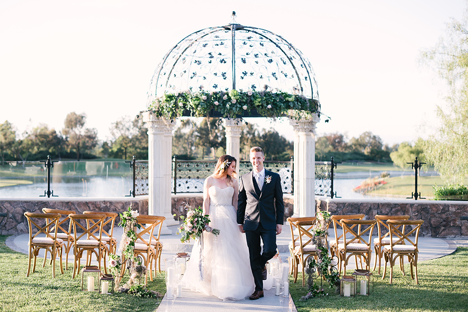 Orange county outdoor wedding at old ranch country club bride strapless ballgown with a sweetheart neckline and beaded detail with groom navy notch lapel suit with matching vest and white dress shirt with long silver tie and white floral boutonniere walking down aisle after ceremony bride holding light pink and green floral bridal bouquet