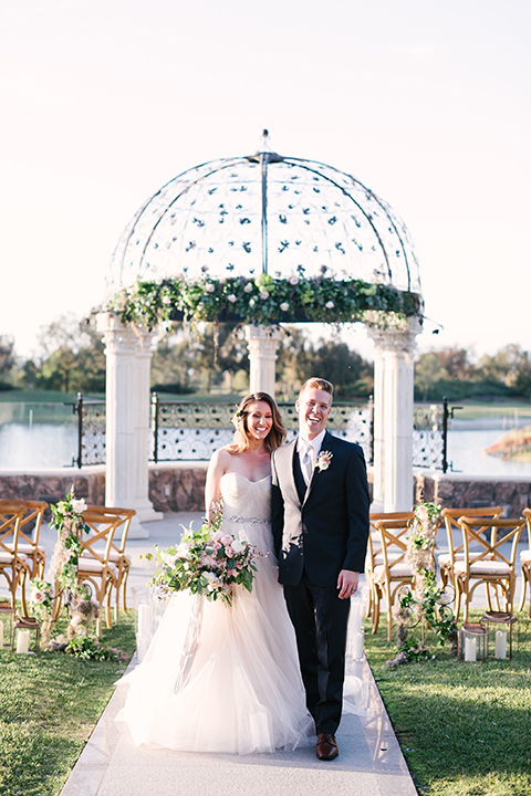 Orange county outdoor wedding at old ranch country club bride strapless ballgown with a sweetheart neckline and beaded detail with groom navy notch lapel suit with matching vest and white dress shirt with long silver tie and white floral boutonniere standing in aisle during ceremony bride holding light pink and green floral bridal bouquet