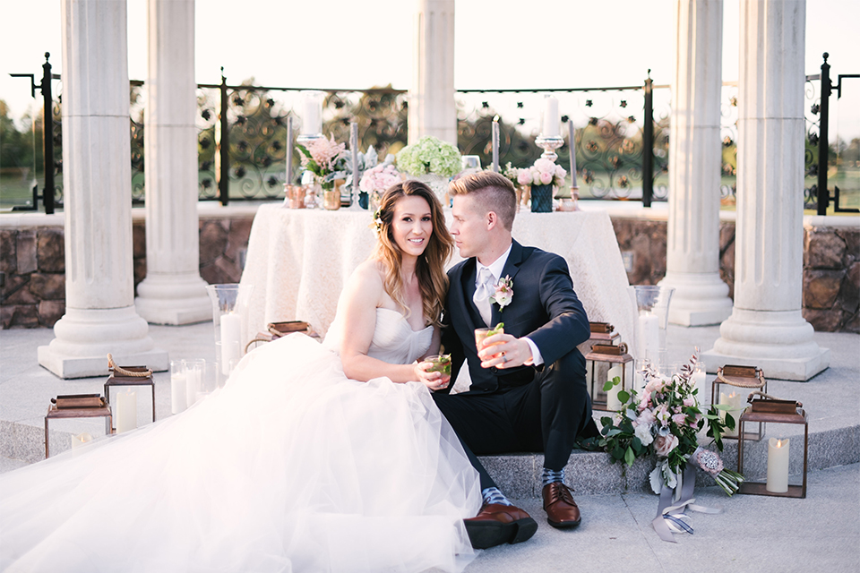 Orange county outdoor wedding at old ranch country club bride strapless ballgown with a sweetheart neckline and beaded detail with groom navy notch lapel suit with matching vest and white dress shirt with long silver tie and white floral boutonniere sitting by table holding drinks