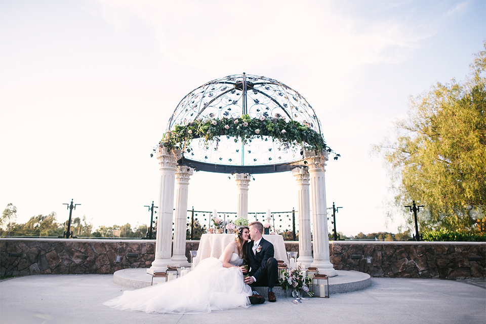 Orange county outdoor wedding at old ranch country club bride strapless ballgown with a sweetheart neckline and beaded detail with groom navy notch lapel suit with matching vest and white dress shirt with long silver tie and white floral boutonniere sitting by table and holding drinks far away