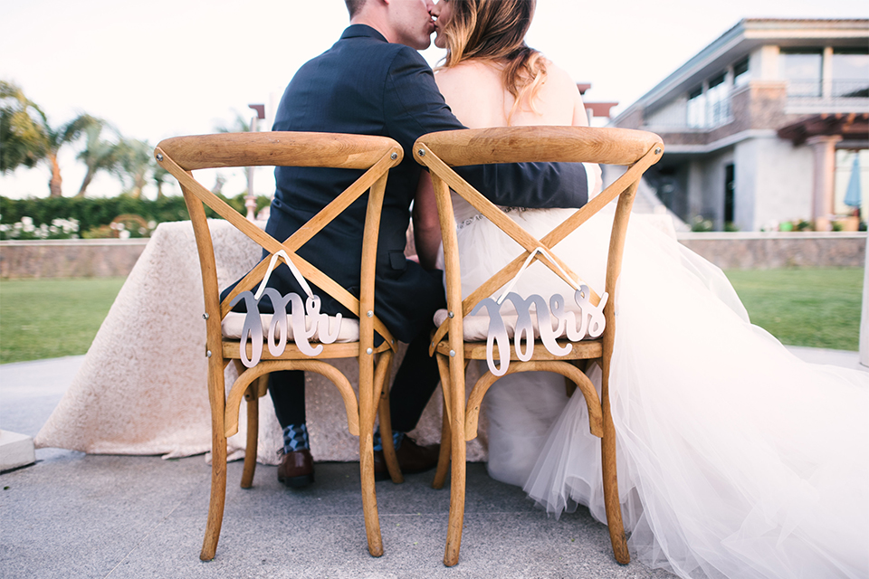 Orange county outdoor wedding at old ranch country club bride strapless ballgown with a sweetheart neckline and beaded detail with groom navy notch lapel suit with matching vest and white dress shirt with long silver tie and white floral boutonniere sitting at table kissing with mr and mrs signs