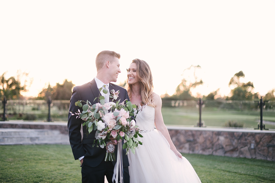 Orange county outdoor wedding at old ranch country club bride strapless ballgown with a sweetheart neckline and beaded detail with groom navy notch lapel suit with matching vest and white dress shirt with long silver tie and white floral boutonniere walking and holding hands bride holding light pink and green floral bridal bouquet