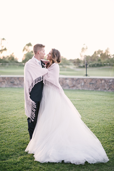 Orange county outdoor wedding at old ranch country club bride strapless ballgown with a sweetheart neckline and beaded detail with groom navy notch lapel suit with matching vest and white dress shirt with long silver tie and white floral boutonniere hugging with blanket wrapped around