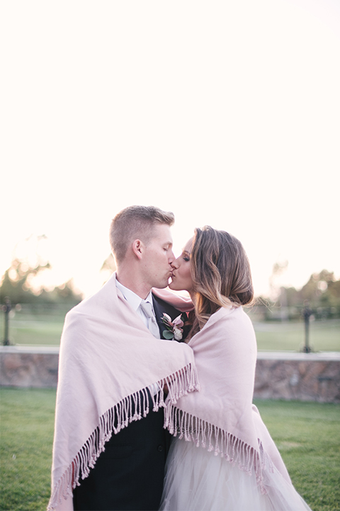 Orange county outdoor wedding at old ranch country club bride strapless ballgown with a sweetheart neckline and beaded detail with groom navy notch lapel suit with matching vest and white dress shirt with long silver tie and white floral boutonniere kissing with blanket wrapped around