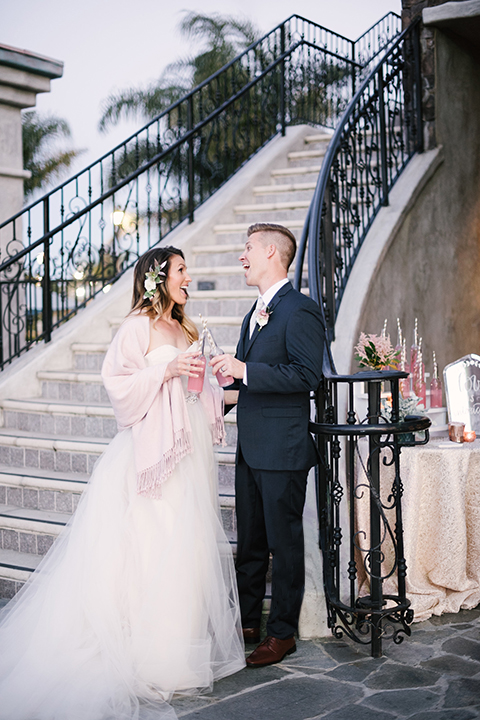 Orange county outdoor wedding at old ranch country club bride strapless ballgown with a sweetheart neckline and beaded detail with groom navy notch lapel suit with matching vest and white dress shirt with long silver tie and white floral boutonniere standing by stairs holding pink drinks