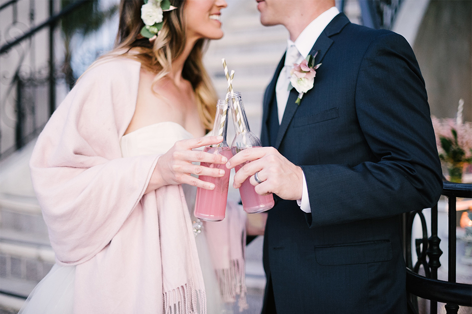 Orange county outdoor wedding at old ranch country club bride strapless ballgown with a sweetheart neckline and beaded detail with groom navy notch lapel suit with matching vest and white dress shirt with long silver tie and white floral boutonniere standing by stairs holding pink drinks