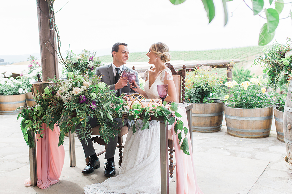 Temecula outdoor wedding at callaway winery bride form fitting lace gown with crystal beading and lace detail with open back design and beaded straps with groom grey notch lapel suit with matching vest and white dress shirt with long black skinny tie and pink floral boutonniere sitting at table holding drinks