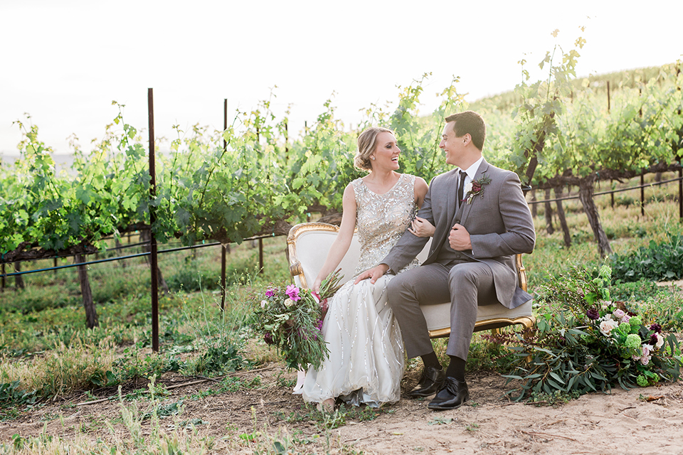 Temecula outdoor wedding at callaway winery bride form fitting lace gown with crystal beading and lace detail with open back design and beaded straps with groom grey notch lapel suit with matching vest and white dress shirt with long black skinny tie and pink floral boutonniere sitting on white vintage couch and bride holding pink and green floral bridal bouquet