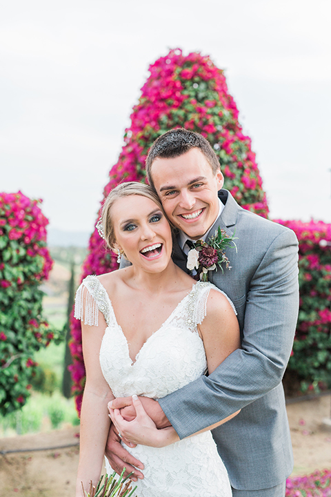 Temecula outdoor wedding at callaway winery bride form fitting lace gown with crystal beading and lace detail with open back design and beaded straps with groom grey notch lapel suit with matching vest and white dress shirt with long black skinny tie and pink floral boutonniere hugging and bride holding pink and green floral bridal bouquet