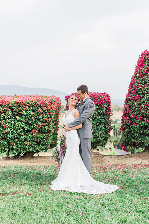 Temecula outdoor wedding at callaway winery bride form fitting lace gown with crystal beading and lace detail with open back design and beaded straps with groom grey notch lapel suit with matching vest and white dress shirt with long black skinny tie and pink floral boutonniere hugging