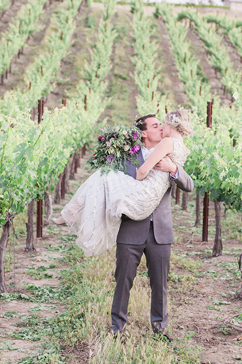 Temecula outdoor wedding at callaway winery bride form fitting lace gown with crystal beading and lace detail with open back design and beaded straps with groom grey notch lapel suit with matching vest and white dress shirt with long black skinny tie and pink floral boutonniere kissing and bride holding pink and green floral bridal bouquet