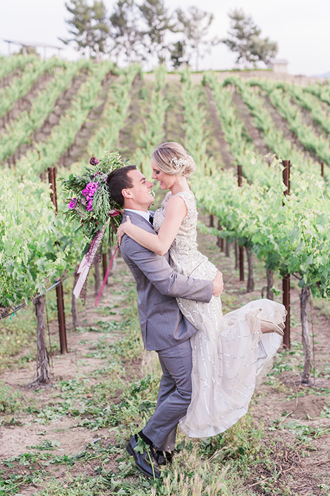 Temecula outdoor wedding at callaway winery bride form fitting lace gown with crystal beading and lace detail with open back design and beaded straps with groom grey notch lapel suit with matching vest and white dress shirt with long black skinny tie and pink floral boutonniere hugging and bride holding pink and green flora bridal bouquet