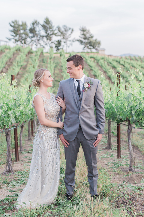 Temecula outdoor wedding at callaway winery bride form fitting lace gown with crystal beading and lace detail with open back design and beaded straps with groom grey notch lapel suit with matching vest and white dress shirt with long black skinny tie and pink floral boutonniere holding hands