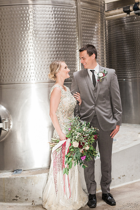 Temecula outdoor wedding at callaway winery bride form fitting lace gown with crystal beading and lace detail with open back design and beaded straps with groom grey notch lapel suit with matching vest and white dress shirt with long black skinny tie and pink floral boutonniere holding hands and bride holding pink and green floral bridal bouquet