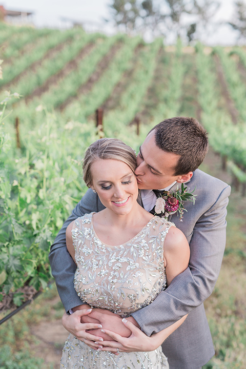 Temecula outdoor wedding at callaway winery bride form fitting lace gown with crystal beading and lace detail with open back design and beaded straps with groom grey notch lapel suit with matching vest and white dress shirt with long black skinny tie and pink floral boutonniere hugging and kissing