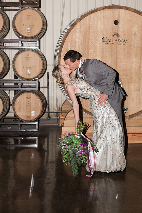 Temecula outdoor wedding at callaway winery bride form fitting lace gown with crystal beading and lace detail with open back design and beaded straps with groom grey notch lapel suit with matching vest and white dress shirt with long black skinny tie and pink floral boutonniere kissing and bride holding pink and green floral bridal bouquet