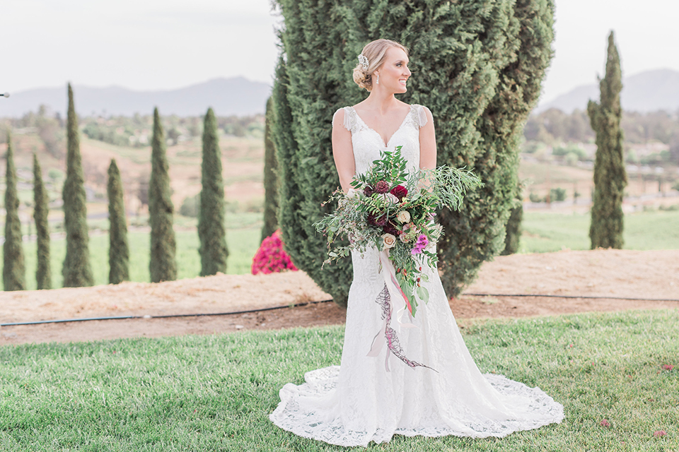 Temecula outdoor wedding at callaway winery bride form fitting lace gown with crystal beading and lace detail with open back design and beaded straps holding pink and green floral bridal bouquet