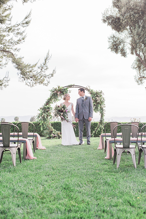 Temecula outdoor wedding at callaway winery bride form fitting lace gown with crystal beading and lace detail with open back design and beaded straps with groom grey notch lapel suit with matching vest and white dress shirt with long black skinny tie and pink floral boutonniere holding hands during cerermony