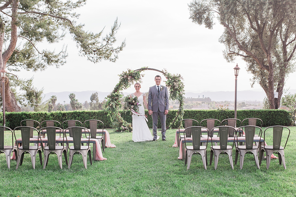 Temecula outdoor wedding at callaway winery bride form fitting lace gown with crystal beading and lace detail with open back design and beaded straps with groom grey notch lapel suit with matching vest and white dress shirt with long black skinny tie and pink floral boutonniere holding hands during ceremony