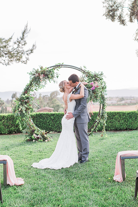 Temecula outdoor wedding at callaway winery bride form fitting lace gown with crystal beading and lace detail with open back design and beaded straps with groom grey notch lapel suit with matching vest and white dress shirt with long black skinny tie and pink floral boutonniere kissing during ceremony