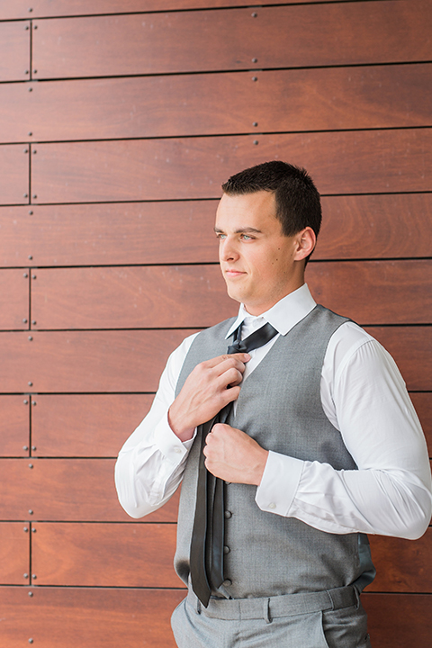 Temecula outdoor wedding at callaway winery groom grey notch lapel suit with matching vest and white dress shirt with long black skinny tie and white and pink floral boutonniere fixing tie getting ready before ceremony