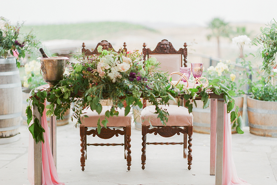 Temecula outdoor wedding at callaway winery table set up light brown wood table with blush pink table runner and white and green flower centerpiece decor with white and gold place settings with blush pink napkins and white and brown vintage chairs