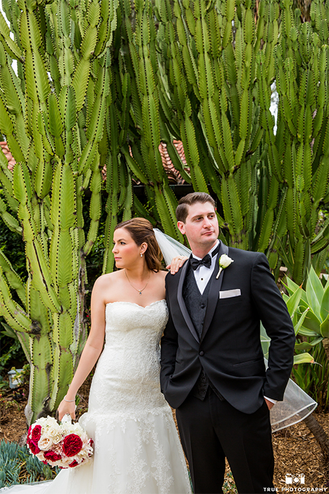 San diego beach wedding at estancia la jolla bride form fitting strapless gown with sweetheart neckline and lace design with long veil and groom black shawl lapel tuxedo with matching vest and white dress shirt with black bow tie and white pocket square with white floral boutonniere hugging and bride holding white and red floral bridal bouquet