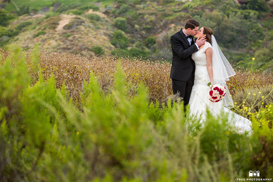 San diego beach wedding at estancia la jolla bride form fitting strapless gown with sweetheart neckline and lace design with long veil and groom black shawl lapel tuxedo with matching vest and white dress shirt with black bow tie and white pocket square with white floral boutonniere kissing and bride holding white and red floral bridal bouquet