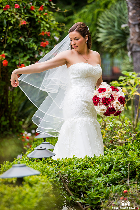 San diego beach wedding at estancia la jolla bride form fitting strapless gown with sweetheart neckline and lace design with long veil holding white and red floral bridal bouquet