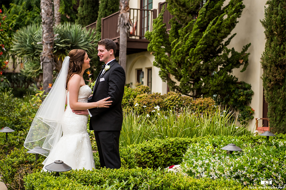 San diego beach wedding at estancia la jolla bride form fitting strapless gown with sweetheart neckline and lace design with long veil and groom black shawl lapel tuxedo with matching vest and white dress shirt with black bow tie and white pocket square with white floral boutonniere hugging