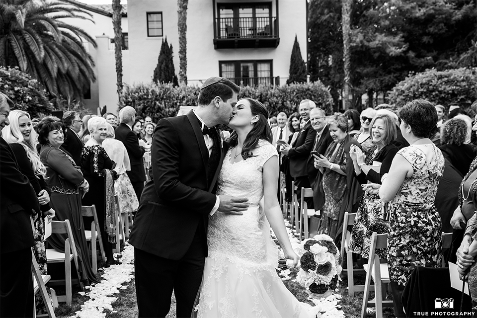 San diego beach wedding at estancia la jolla bride form fitting strapless gown with sweetheart neckline and lace design with long veil and groom black shawl lapel tuxedo with matching vest and white dress shirt with black bow tie and white pocket square with white floral boutonniere kissing after ceremony black and white photo