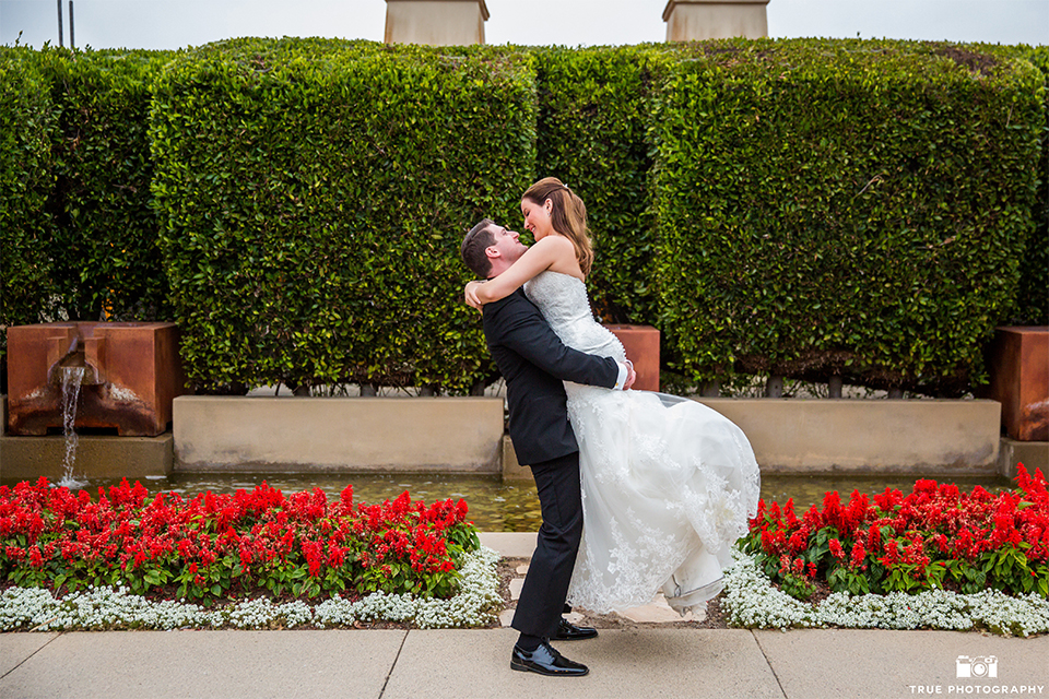 San diego beach wedding at estancia la jolla bride form fitting strapless gown with sweetheart neckline and lace design with long veil and groom black shawl lapel tuxedo with matching vest and white dress shirt with black bow tie and white pocket square with white floral boutonniere hugging