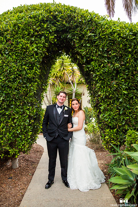 San diego beach wedding at estancia la jolla bride form fitting strapless gown with sweetheart neckline and lace design with long veil and groom black shawl lapel tuxedo with matching vest and white dress shirt with black bow tie and white pocket square with white floral boutonniere holding hands