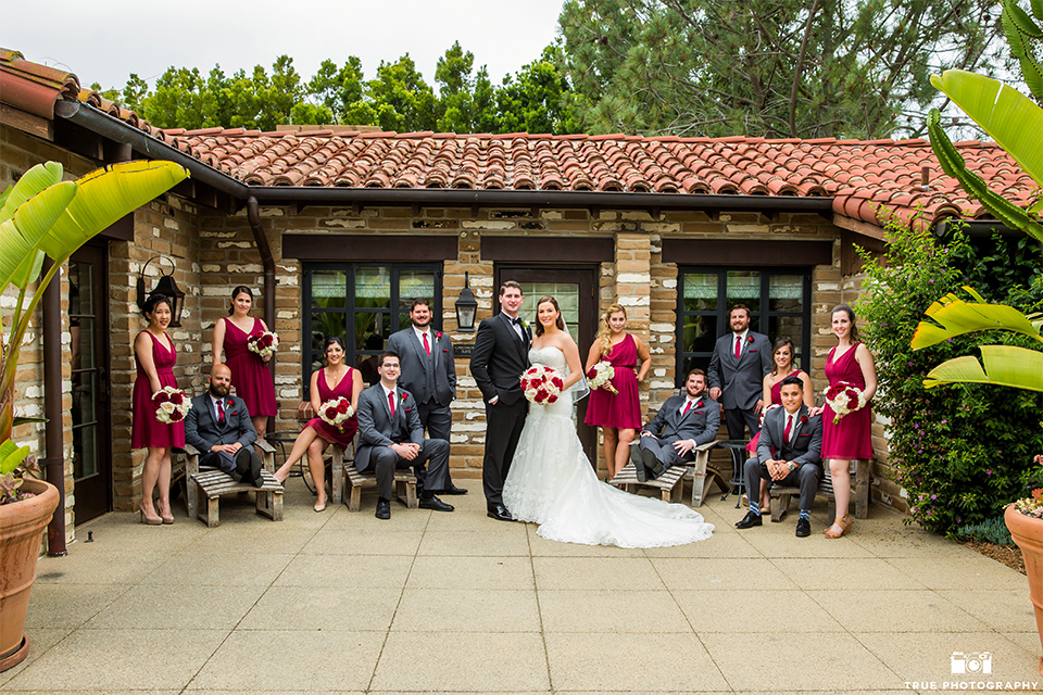 San diego beach wedding at estancia la jolla bride form fitting strapless gown with sweetheart neckline and lace design with long veil and groom black shawl lapel tuxedo with matching vest and white dress shirt with black bow tie and white pocket square with white floral boutonniere with wedding party bridesmaids short red dresses and groomsmen charcoal grey suits with long red ties