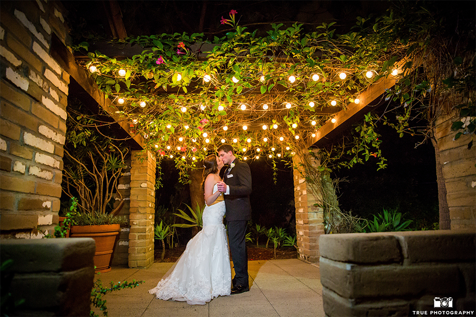 San diego beach wedding at estancia la jolla bride form fitting strapless gown with sweetheart neckline and lace design with long veil and groom black shawl lapel tuxedo with matching vest and white dress shirt with black bow tie and white pocket square with white floral boutonniere dancing under lights