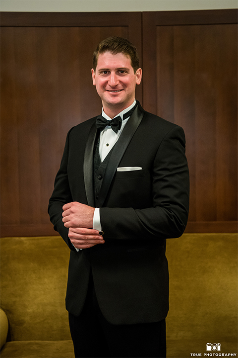 San diego beach wedding at estancia la jolla groom black shawl lapel tuxedo with black bow tie and white dress shirt with white pocket square and white floral boutonniere holding sleeve