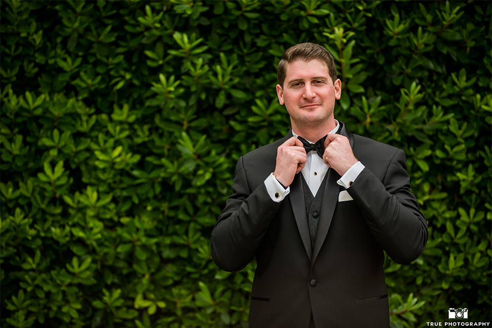 San diego beach wedding at estancia la jolla groom black shawl lapel tuxedo with black bow tie and white dress shirt with white pocket square and white floral boutonniere fixing tie