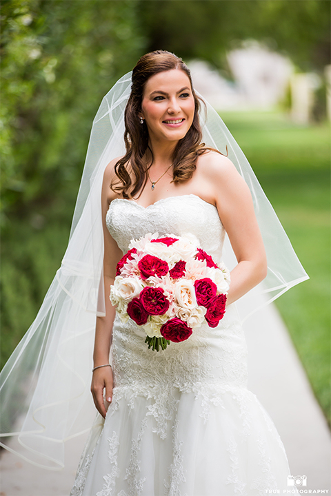 San diego beach wedding at estancia la jolla bride form fitting strapless gown with sweetheart neckline and lace design with long veil holding white and red floral bridal bouquet