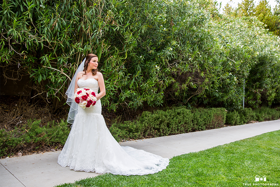 San diego beach wedding at estancia la jolla bride form fitting strapless gown with sweetheart neckline and lace design with long veil holding white and red floral bridal bouquet