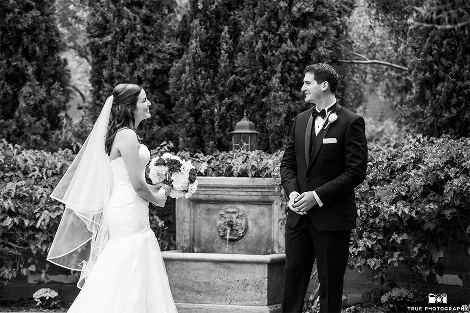 San diego beach wedding at estancia la jolla bride form fitting strapless gown with sweetheart neckline and lace design with long veil and groom black shawl lapel tuxedo with matching vest and white dress shirt with black bow tie and white pocket square with white floral boutonniere first look black and white photo