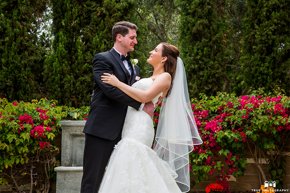 San diego beach wedding at estancia la jolla bride form fitting strapless gown with sweetheart neckline and lace design with long veil and groom black shawl lapel tuxedo with matching vest and white dress shirt with black bow tie and white pocket square with white floral boutonniere hugging 