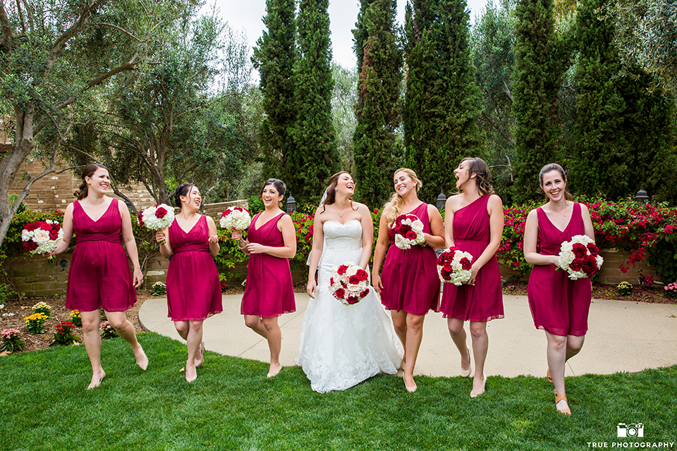 San diego beach wedding at estancia la jolla bride form fitting strapless gown with sweetheart neckline and lace design with long veil holding white and red floral bridal bouquet with bridesmaids short red dresses holding white and red floral bridal bouquets walking