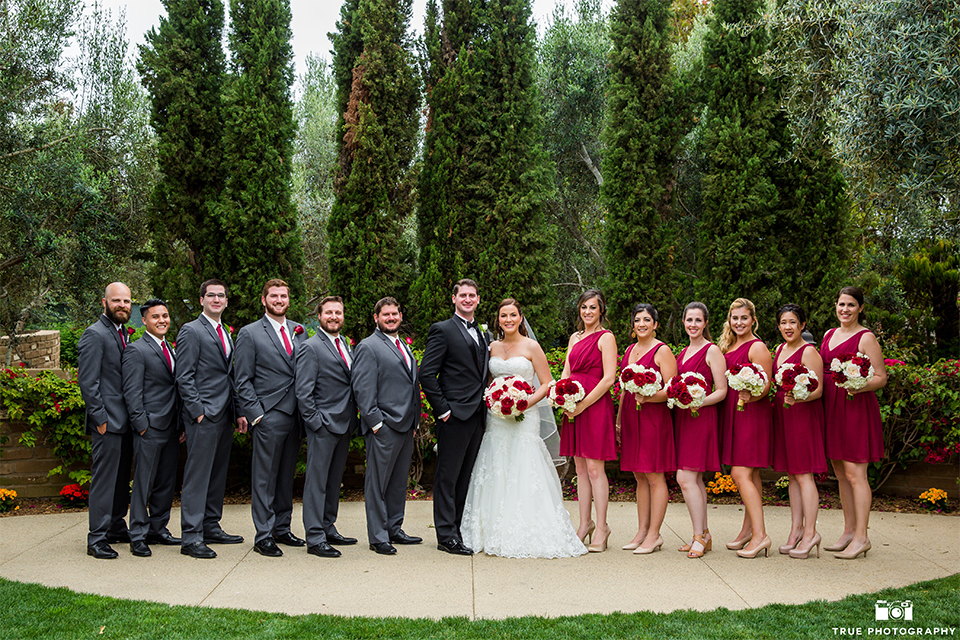 San diego beach wedding at estancia la jolla bride form fitting strapless gown with sweetheart neckline and lace design with long veil and groom black shawl lapel tuxedo with matching vest and white dress shirt with black bow tie and white pocket square with white floral boutonniere with bridesmaids short red dresses and white and red floral bouquets and groomsmen charcoal grey suits with long red ties